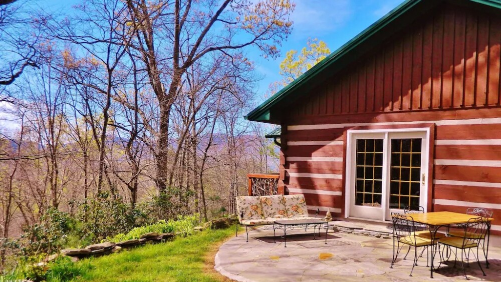 A woodsy cabin with outside patio furniture, and the woods and mountain in the background.