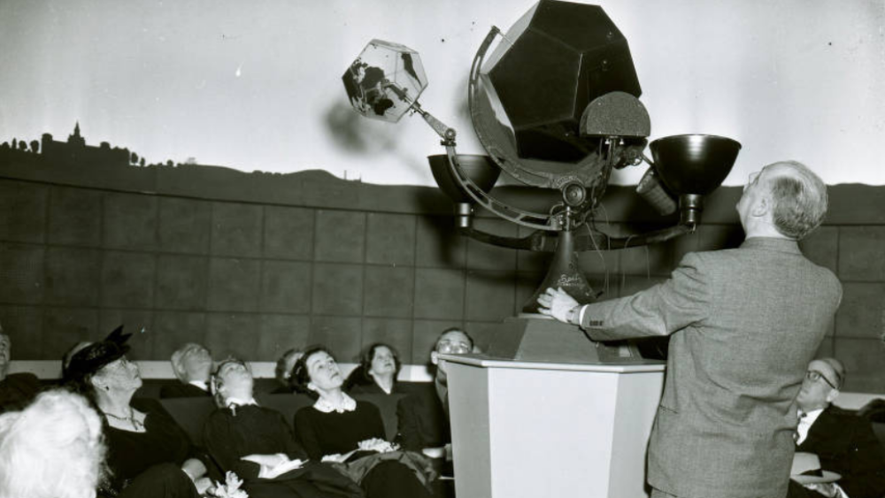 Someone stands in front of a tool sitting on a pedestal as a circle of people sit around it in a black and white photograph.