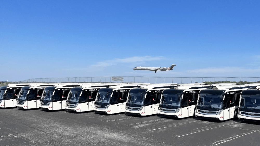 A plane flies over a fleet of shuttle buses parked in a lot.