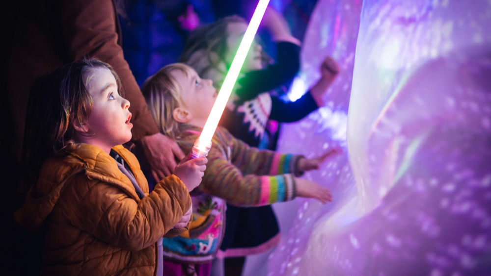 A small child stares starry eyed at a light saber they hold in their hands, while other kids touch a glowing squishy looking art installation at the Portland Winter Light Festival.