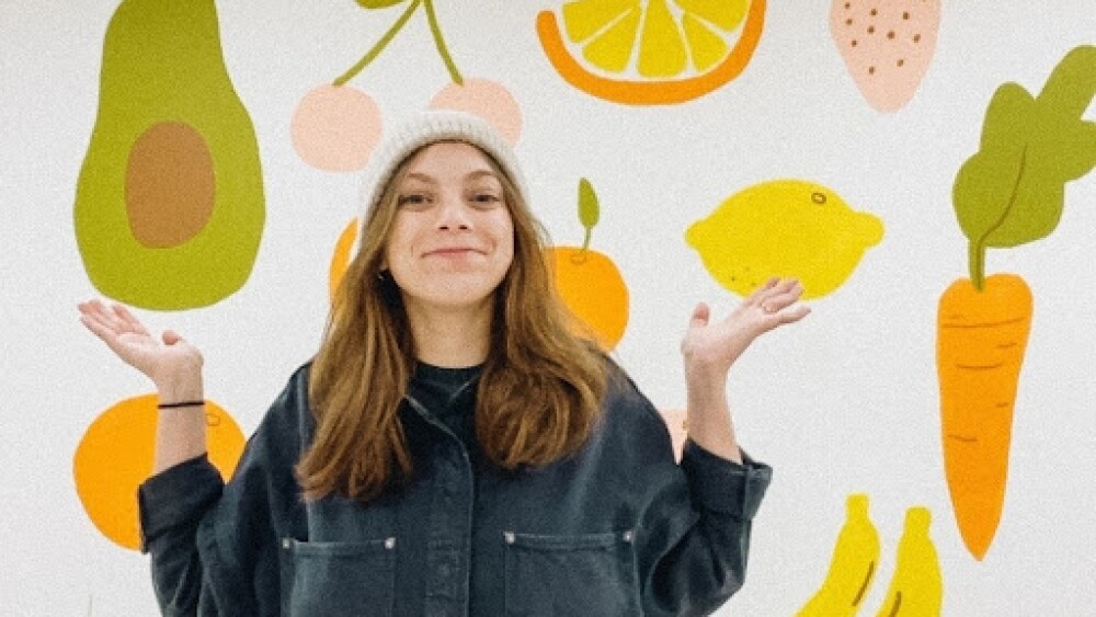 A smiling artist wearing a beanie and oversized button-up raises her palms in front of a mural of fruit including an avocado, cherries, orange slice, lemon, carrot, and strawberry.