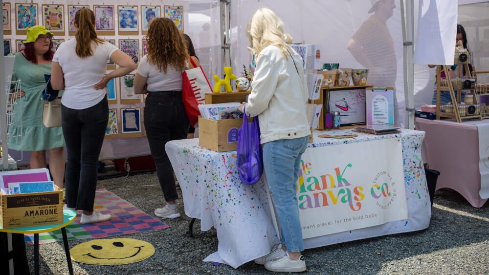 A shopper with a purple shopping bag stands in front of a tented booth that features artistic prints. A sign on the booth's table reads "The Blank Canvas Co." Inside of the booth, four more people stand around talking.