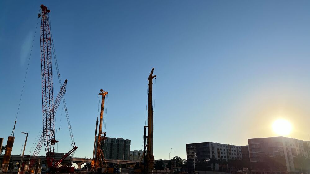 The construction site at The Pendry in downtown Tampa. The sun is setting over the riverfront apartments in the distance, with cranes and machinery in the left foreground of the picture behind protective construction fences.