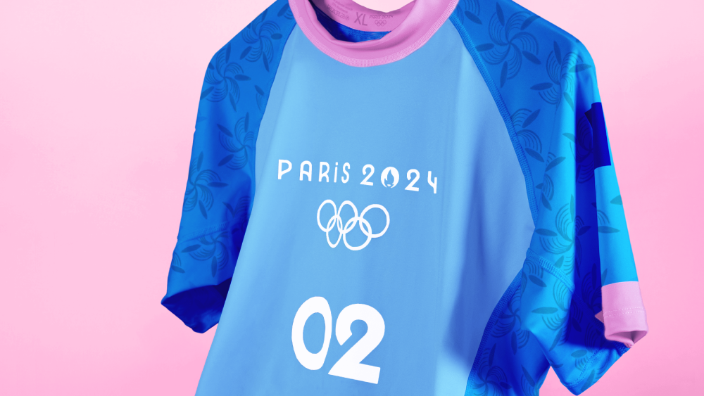 A light blue and dark blue rash guard top that will be used as the official uniform of Olympians competing at the Paris 2024 Olympics. It has short sleeves, pink trim on the sleeves and collar, and a flower-like design on the dark blue portion along the sides.