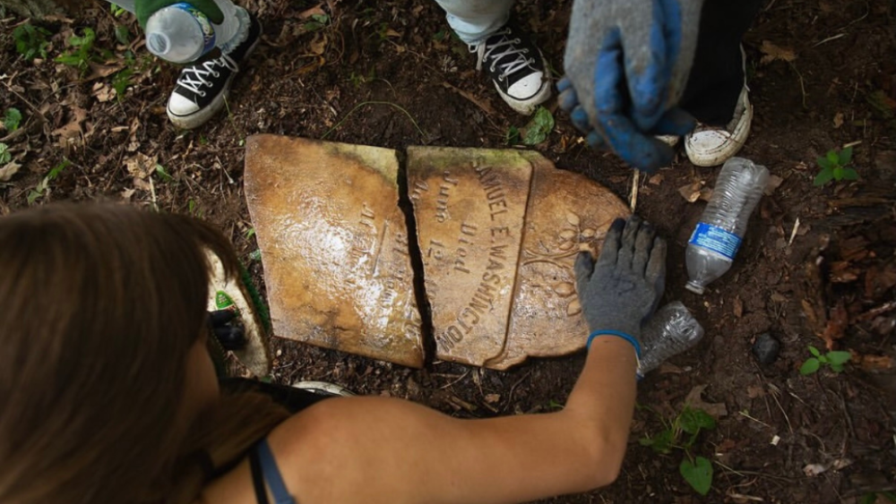 A birds-eye style photo depicting two people, partially in frame, leaning over a gravestone; The gravestone is broken and lays flat on the ground; there are two water bottles in the photo; one person holds a gloved hand near the top of the gravestone.