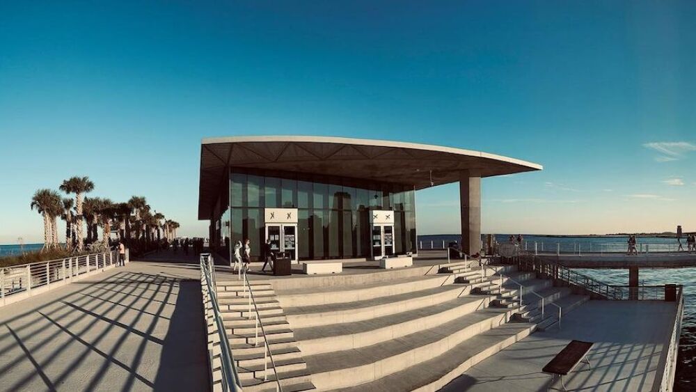 The St. Pete Pier surrounded by water with blue skies above. There are some people walking around and palm trees lining the walkway.