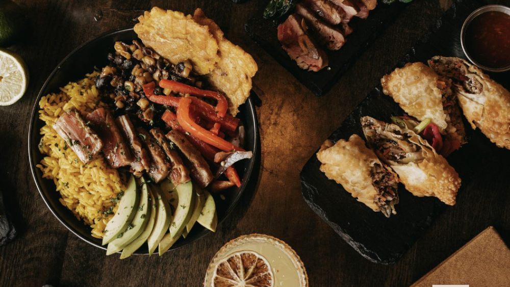 A table with multiple plates of food, including steak, seared ahi tuna, and chicken wings. There is also a cocktail or mocktail and moody lighting.