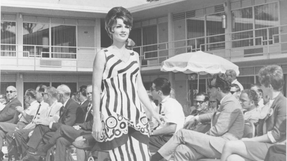 A black-and-white photo of a model in a striped dress on a catwalk in front of a seated audience in 1969.