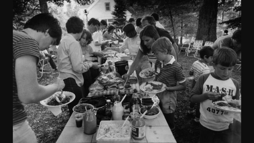 Black and white photograph of Gayton Kirk members and members of visiting Scots and Massachusetts groups getting food during outdoor picnic; they file along buffet table and serve food to themselves.