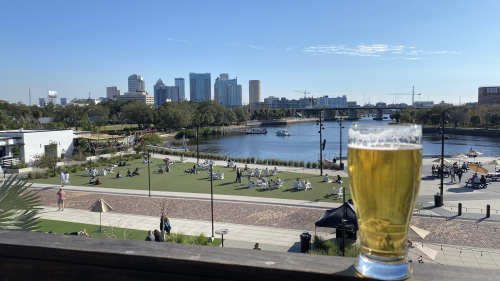 A beer sits on a ledge at Armature Works. The Hillsborough River sits behind the glass, and Tampa's skyline is visible in the background. People are by the river, and the sky barely has a cloud in it.
