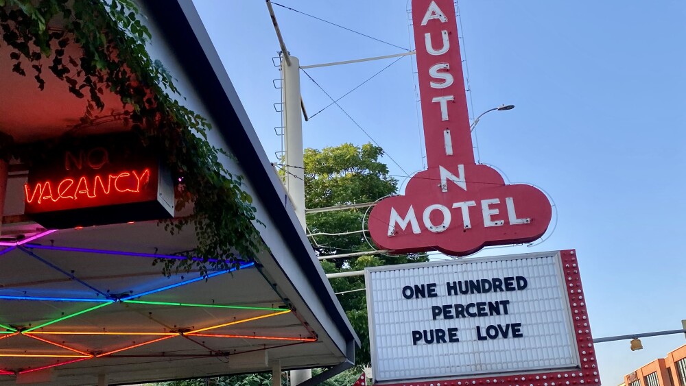 A view of the iconic Austin Motel sign, which is shaped like a large, red, vertical arrow pointing downward. The sign reads 'AUSTIN MOTEL' in white letters and below it, a marquee displays the message 'ONE HUNDRED PERCENT PURE LOVE.' To the left, there is a 'No Vacancy' sign glowing in red neon under a canopy adorned with colorful lights. The canopy is covered in lush greenery, adding a vibrant touch to the scene. The clear blue sky in the background enhances the bright and welcoming atmosphere of this well-known Austin landmark.