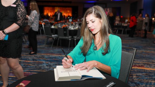 An author autographs her book in a ballroom with tables and dishes in the background.