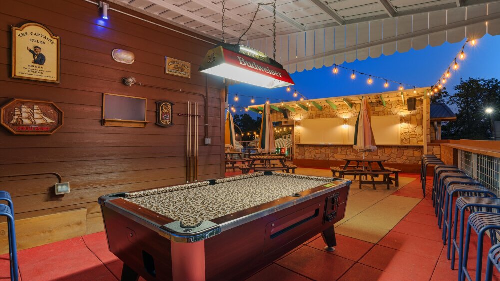 A pool table with a leopard print cover, a retro lamp reading "Budweiser" overhead, with a chalkboard, cues, and decor on the back wall. In the background, there's an outdoor patio with umbrella-covered picnic tables.
