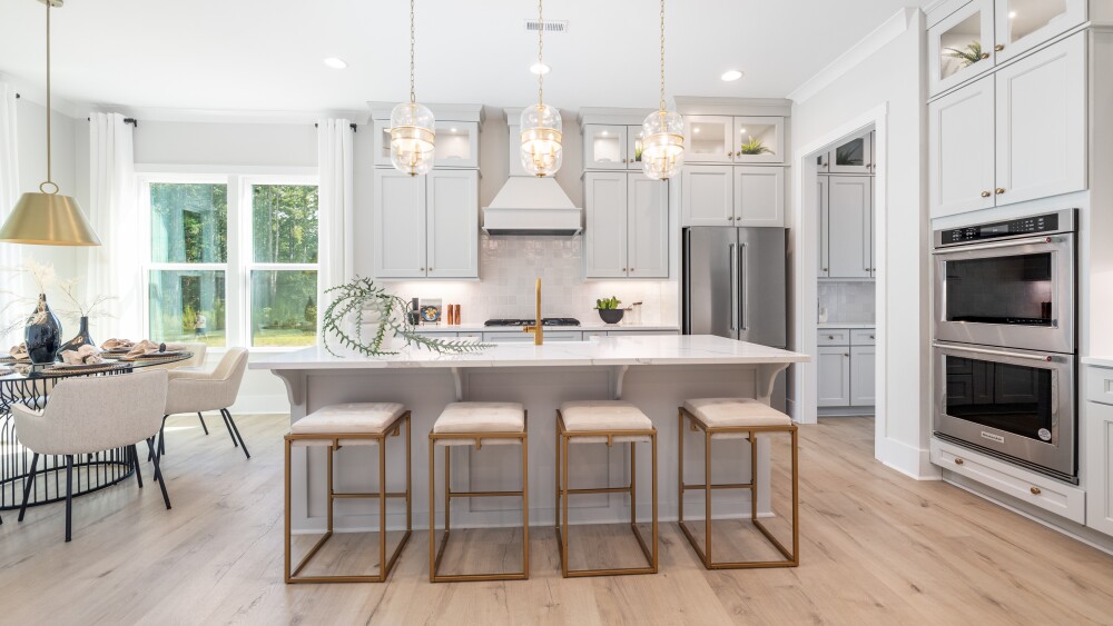 The interior of a new home, with the camera facing the kitchen and stone island. Four gold stools are arranged in front of the island.