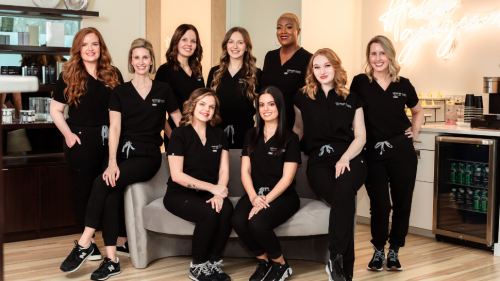 A group of nine people posing around a couch, all smiling at the camera while wearing black scrubs.