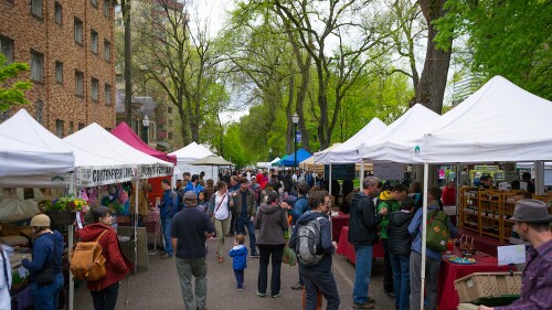 People walk down an aisle at the Portland Farmers Market, where canopies and vendors are set up on either side.