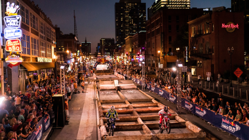 Two people on dirt bikes drive over a series of logs covering Lower Broadway as people watch on the sidewalk under neon signs.