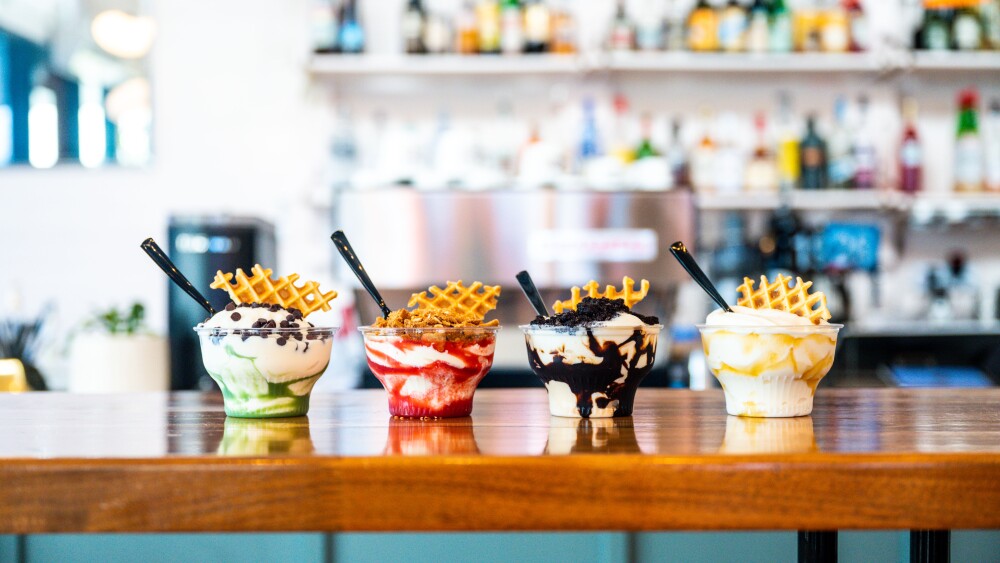 Four different flavors of custard lined up on a wooden counter, topped with waffles and various toppings.