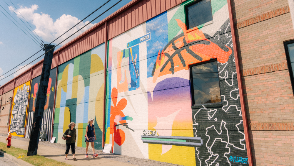 Multiple large-scale murals on the side of a building with two people walking in front of them at Artville.