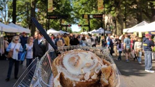 A clear plastic container holding a large cinnamon bun with white frosting on top. In the background, many people are standing or walking on a tree-lined street.