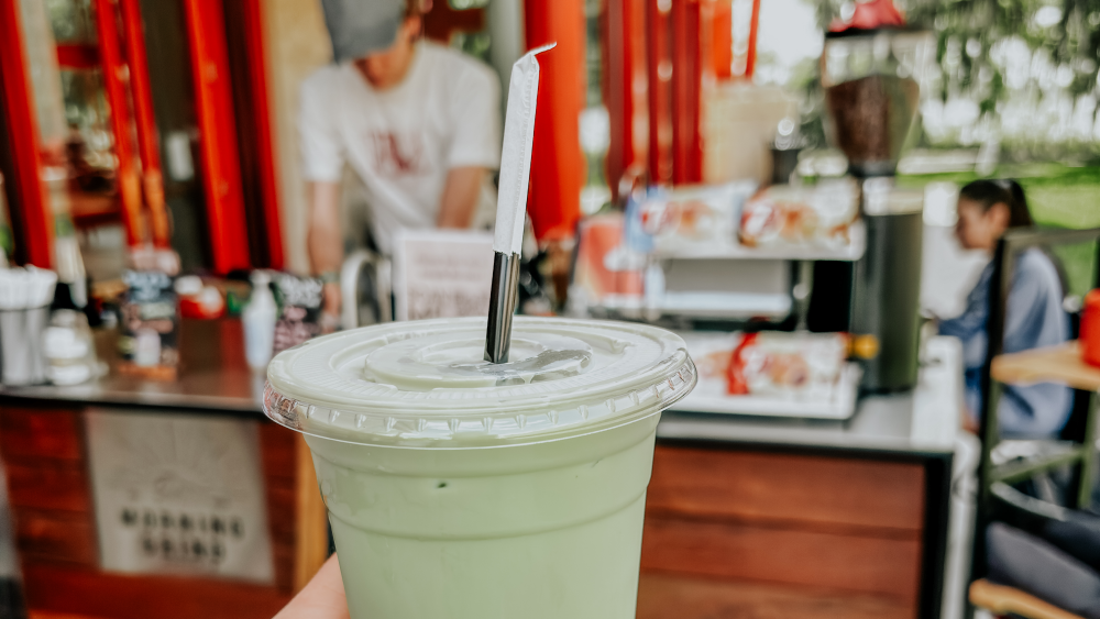 A hand holds a green colored iced coffee in a plastic cup in the foreground. In the background is a blurry shot of students working the Morning Grind coffee cart.