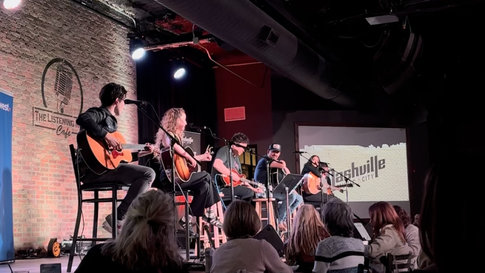 Five songwriters, each with a guitar in hand, sitting in tall chairs with microphone stands in front of each.