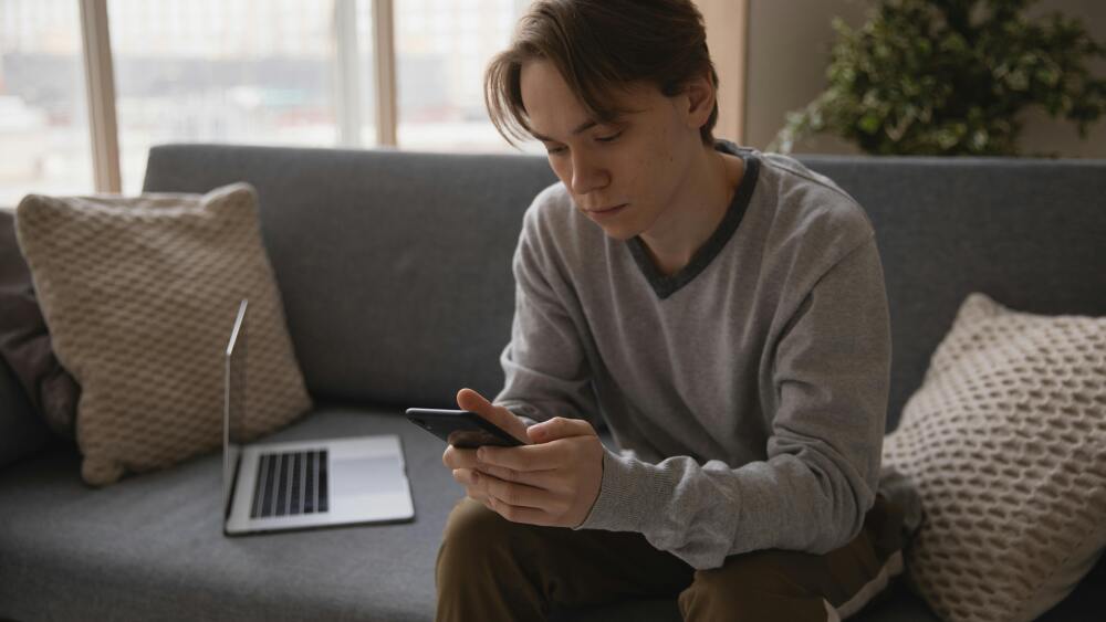 A boy sits on a couch looking at his smart phone, with a laptop next to him.