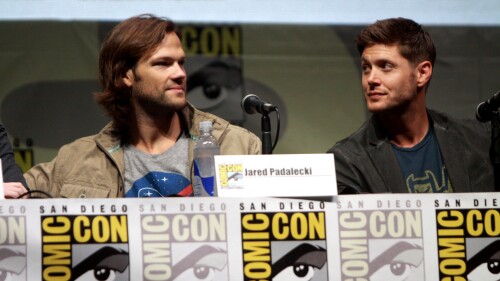 Jared Padalecki and Jensen Ackles sit at a Comic-Con panel. Padalecki is on the left with shoulder-length hair and a casual jacket over a graphic t-shirt, with a name card reading "Jared Padalecki" in front of him. Ackles is on the right, with shorter hair and wearing a dark jacket, looking toward him. Both are seated behind microphones, with Comic-Con logos on the table and the backdrop behind them. Bottled water is on the table.