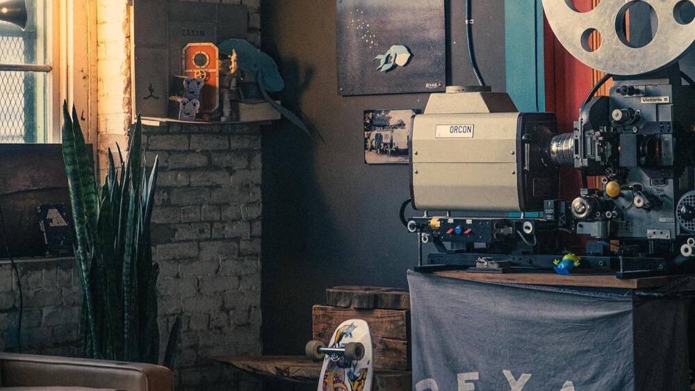 A vintage 35mm projector in a brick room on top of a stand with a Bexar TX flag below it.