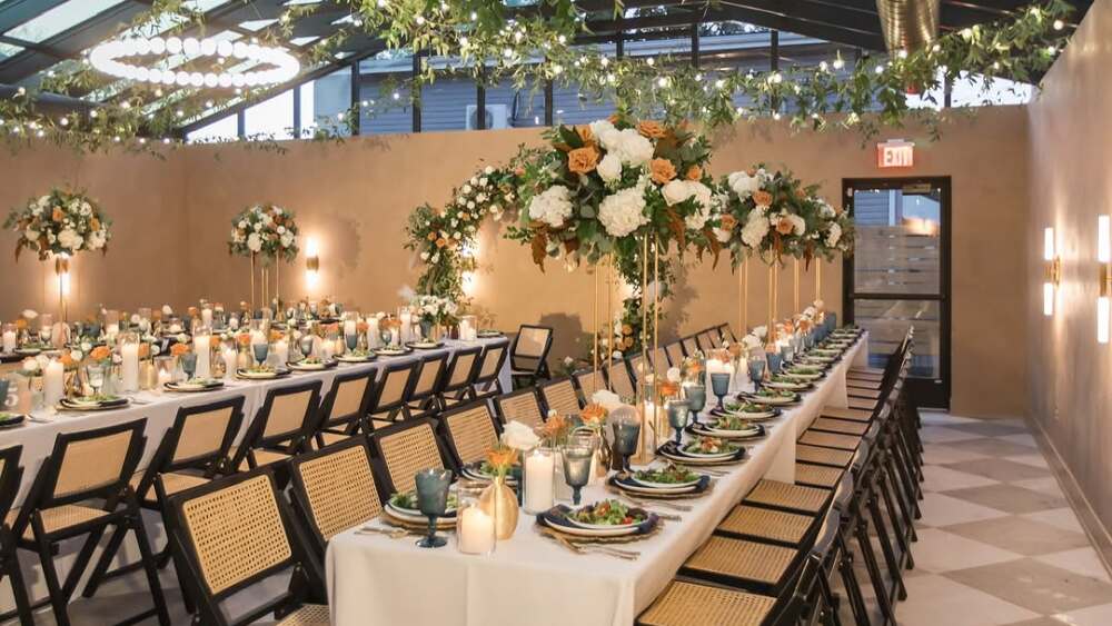 An atrium style room with marble checkered floor set up for a wedding with rows of tables featuring white linens, peach and white flowers, candles, and greenery and lights hanging from the ceiling.