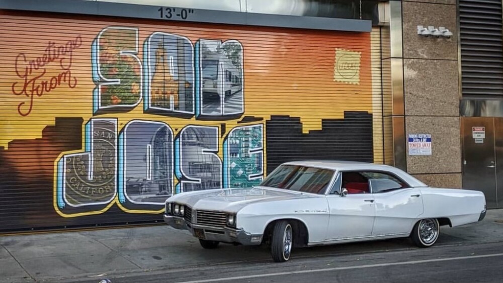 Ricardo Cortez's white lowriding car, in front of a mural on the side of a building that says "Greetings from San Jose" with a stylized city skyline.