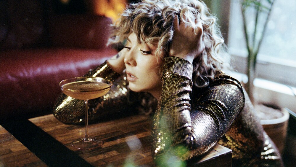 A woman wearing a glittery top, staring into a cocktail in front of her.