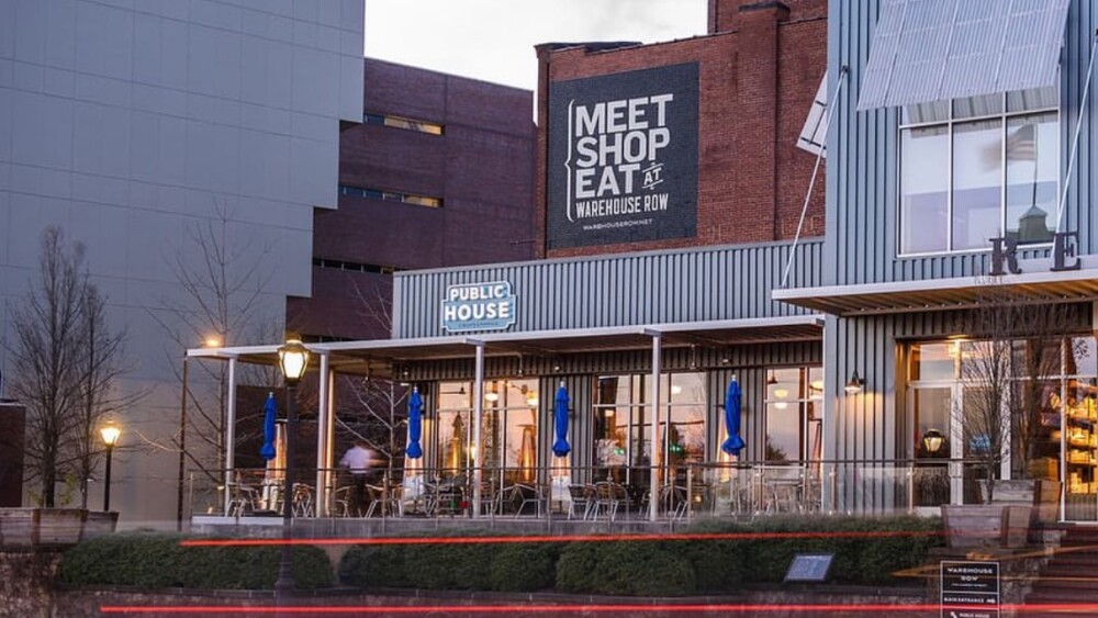 Street view of a modern restaurant named "Public House" at dusk, featuring outdoor seating with blue umbrellas. Nearby sign reads "Meet Shop Eat." Tail lights streak by, adding motion.