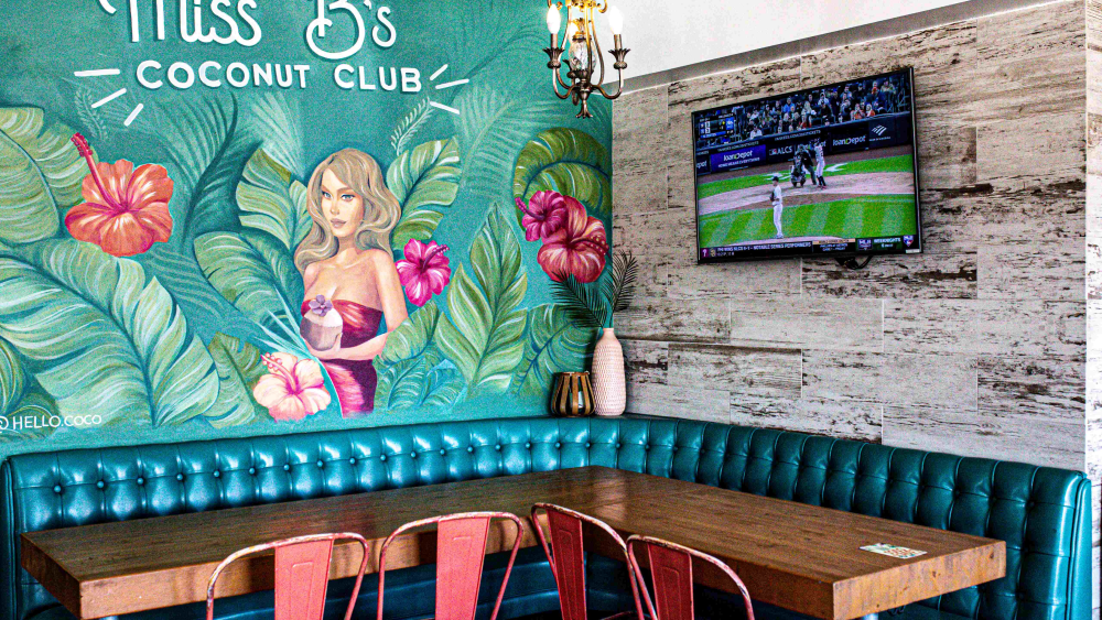 The interior of Miss B's Coconut Club in San Diego; one wall is painted with a tropical mural featuring green leaves, pink and orange hibiscus flowers, and a blonde woman holding a coconut drink.