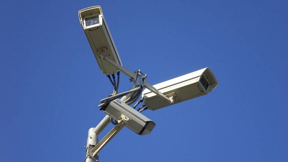 Three white speeding cameras on a pole, pointed in three different directions, set against a blue sky.