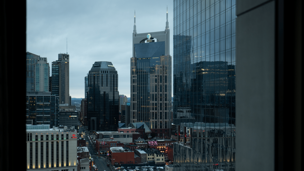 A view of a moody downtown skyline in darker hues of blue from a skyscraper's window. You can make out Broadway, the We Work building, and the Batman Building.