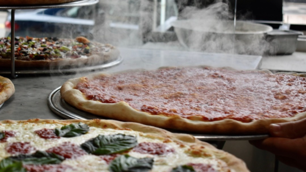 Multiple pizzas sit on plates with steam coming off the top. They are on a silver countertop with five pies visible, with several different flavor combinations visible.