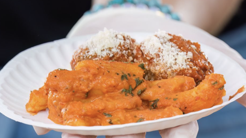 A paper plate of paste and meatballs is being held toward the camera by a gloved hand. It is close up, and there is red sauce + parmesan cheese atop the dish.