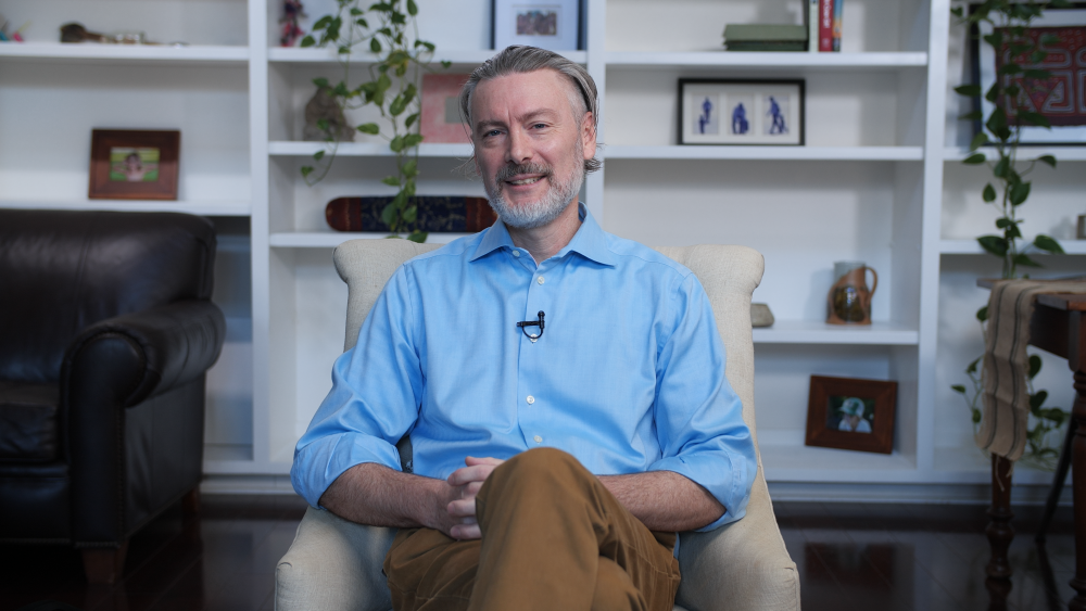 a man sits in a chair smiling with his legs crossed