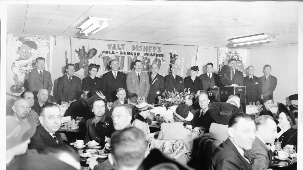 Disney and associates stand under a banner reading "Walt Disney's Full-Length Feature" at a business luncheon.