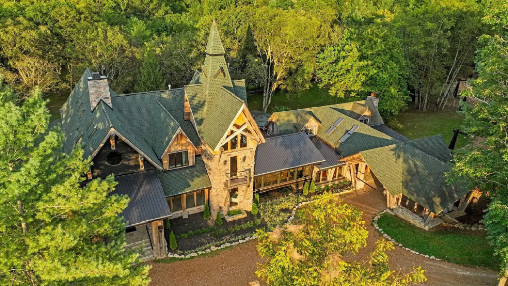 Aerial view of a lodge-style estate with stone accents, green roofs, and a central tower surrounded by trees.