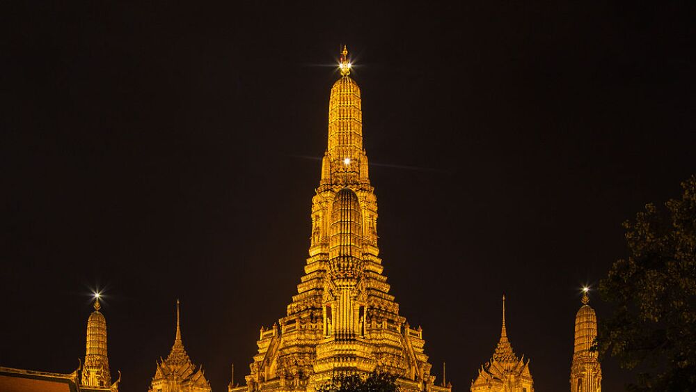 The towering spires of an orange temple rise majestically against the black sky. The intricate details of the structure are highlighted, and the temple’s distinctive design is visible, featuring a central prang (tower) surrounded by smaller towers.