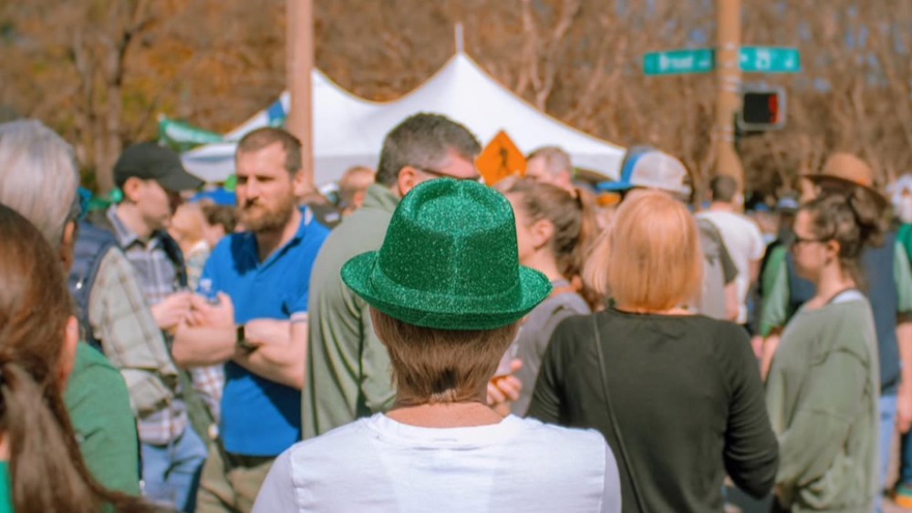 A person wearing a glittery green fedora stands, faced away, in front of a crowd of people. A white tent and bare trees are seen in the background.
