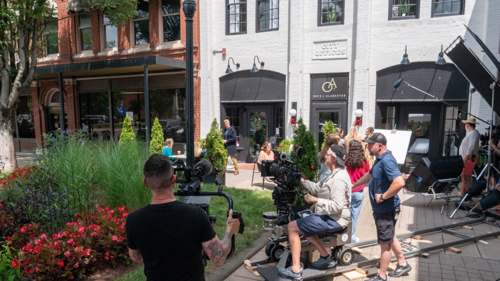 A holiday movie being filmed outdoors in downtown Franklin, TN, with production crew members holding cameras and mics. Some trees in the background are decorated with ornaments.