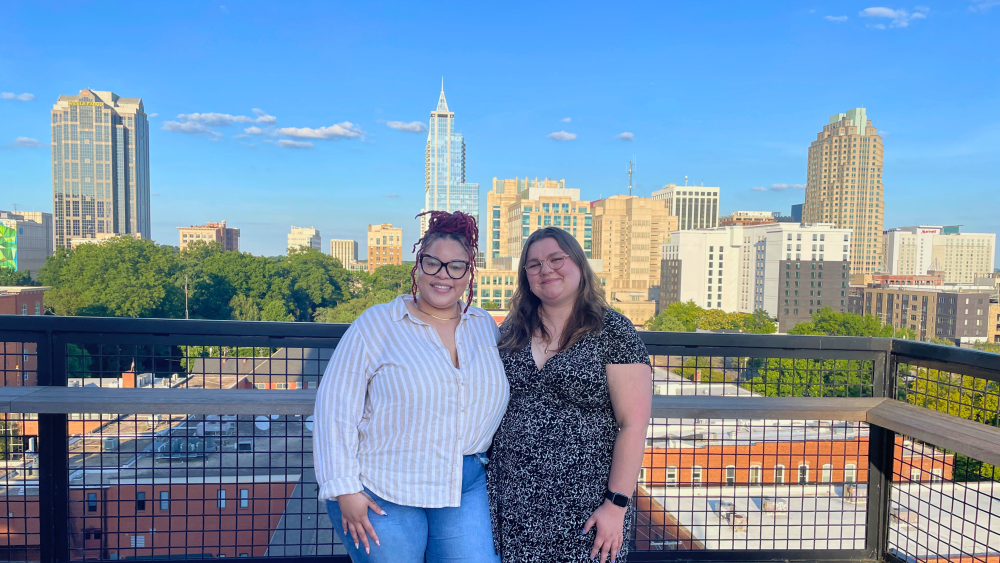 Two women stand on a rooftop in front of Raleigh's skyline