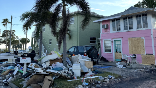A pile of debris outside a pink home in Gulfport. The home's windows are boarded up following Hurricane Helene, and there is standing water in the street. There are clear skies above, and it looks like the sun is setting at dusk.