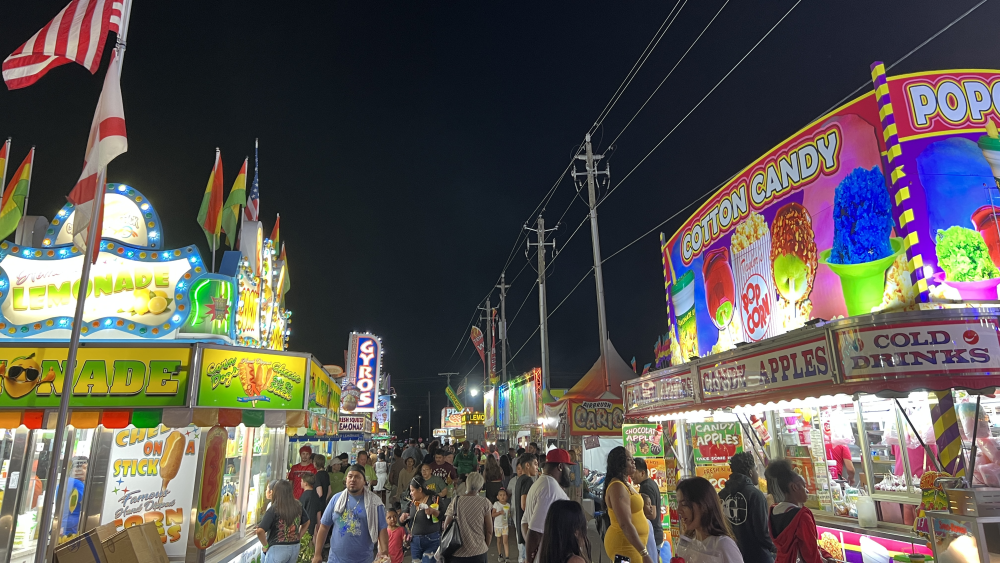 The Florida Strawberry Festival at night. Crowds walk between colorful, lit booths selling food like candy apples, lemonade, corndogs, and cotton candy.