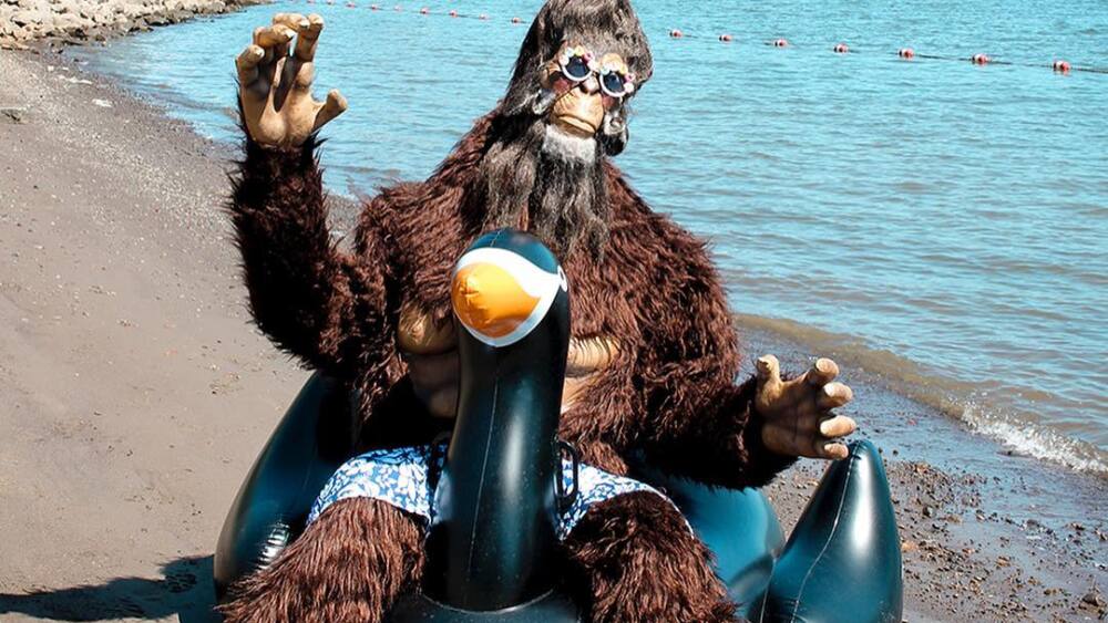 A person in a sasquatch suit sits on an inflatable bird floaty along the bank of a river on a sunny day with a city skyline in the background. 