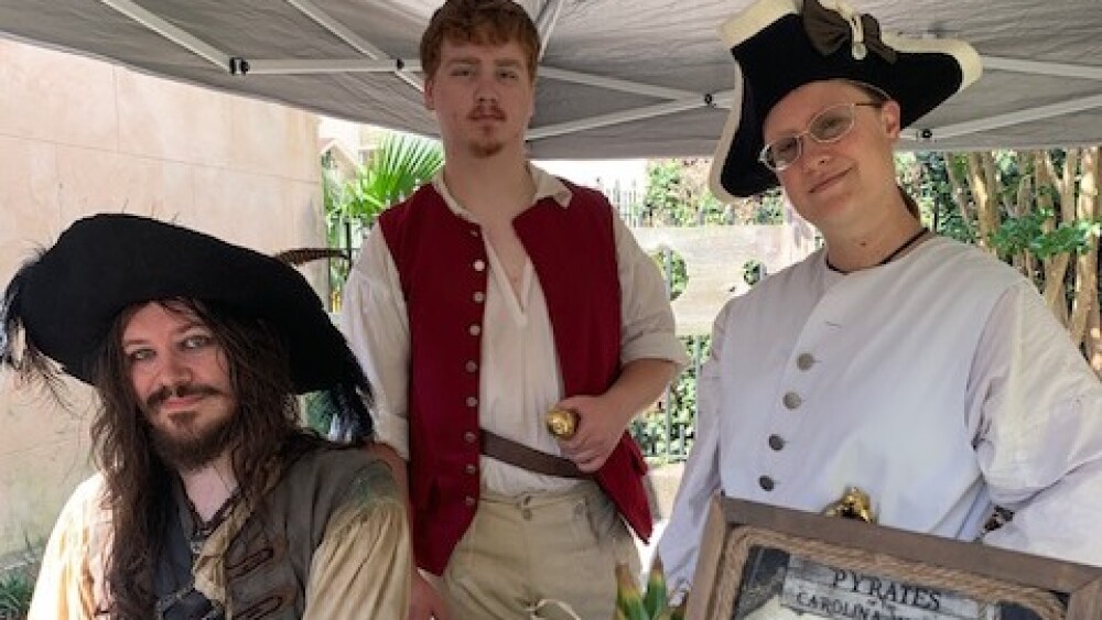 three pirate reenactors pose for the camera. The pirates have on pirate gear white shirts with red, white and brown vests and props like a sword, a cup, a picture frame.
