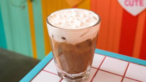 An iced almond mocha in a clear glass topped with toasted marshmallow and mini marshmallows with a colorful wall. out of focus in the background.